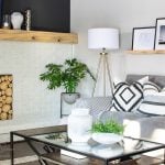 A modern family room showcases a white brick fireplace with a stack of logs, complemented by a gray sectional sofa and patterned cushions. A tripod floor lamp stands beside a glass coffee table, while framed art and a potted plant reveal the charm on a wooden shelf.
