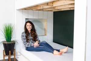 A woman with long hair, wearing a black and white checkered shirt and jeans, sits on a white mattress inside a partially built wooden structure with built-in features. To her left, against a white wall, stands a tall potted plant.