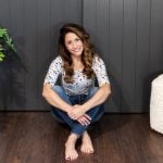 A woman with long, wavy hair sits on a wooden floor, leaning against a dark gray paneled wall she transformed during her DIY closet door makeover. She's wearing a patterned top and jeans, smiling, with her hands crossed in front. A decorative plant and a cushioned chair are nearby.