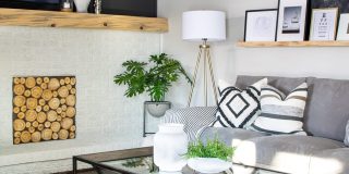 A modern family room showcases a white brick fireplace with a stack of logs, complemented by a gray sectional sofa and patterned cushions. A tripod floor lamp stands beside a glass coffee table, while framed art and a potted plant reveal the charm on a wooden shelf.