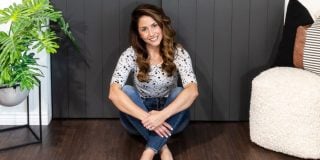 A woman with long, wavy hair sits on a wooden floor, leaning against a dark gray paneled wall she transformed during her DIY closet door makeover. She's wearing a patterned top and jeans, smiling, with her hands crossed in front. A decorative plant and a cushioned chair are nearby.